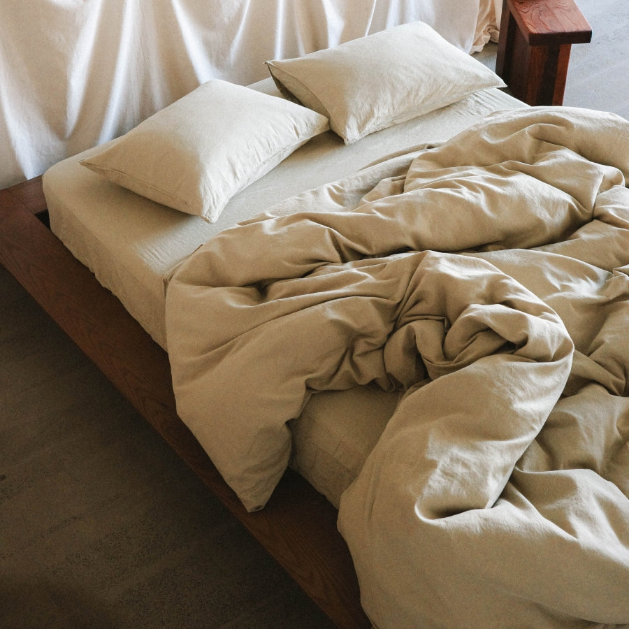 Natural light green bedding set with a crumpled duvet cover and two matching pillows on a dark wooden bed frame.