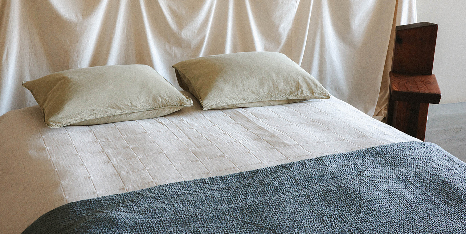 Bed with light green pillows, white quilt and a blue blanket against a white curtain background