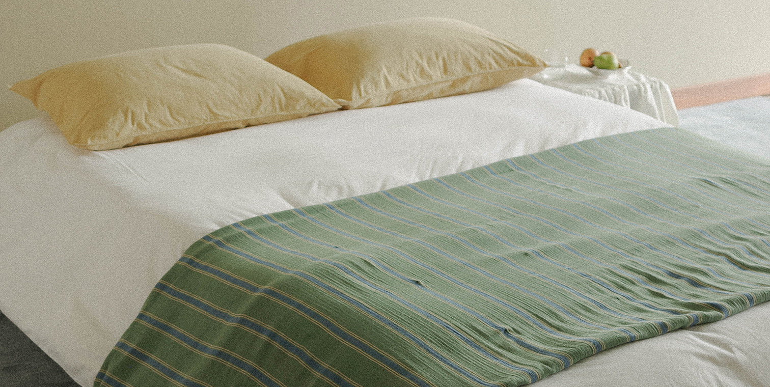 Bed with green striped blanket and yellow pillows and white duvet on a neutral background
