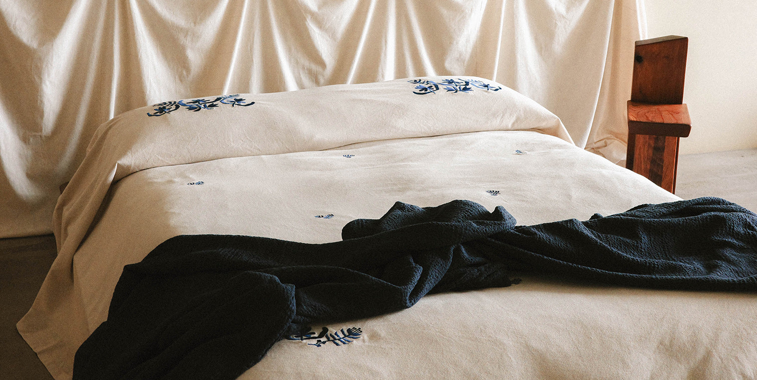 Beige bedspread with blue floral patterns on a bed, with a dark blue blanket draped over it.
