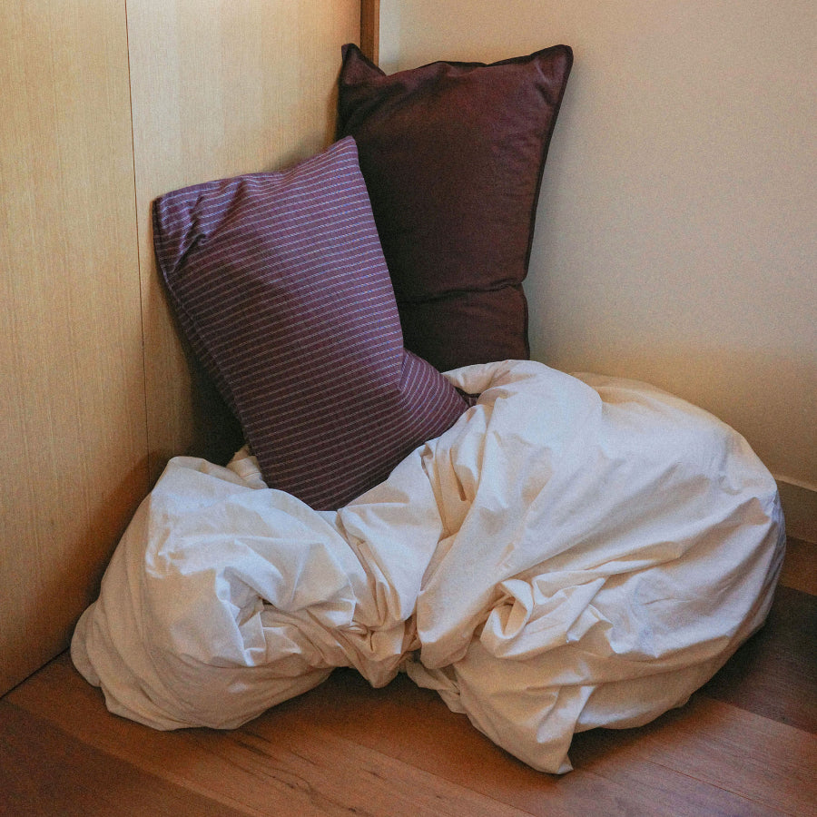 A rumpled white duvet sits on a wooden floor in the corner of a room, with two pillows leaning against the wall: one dark plum pillow and one striped mauve-purple pillow.