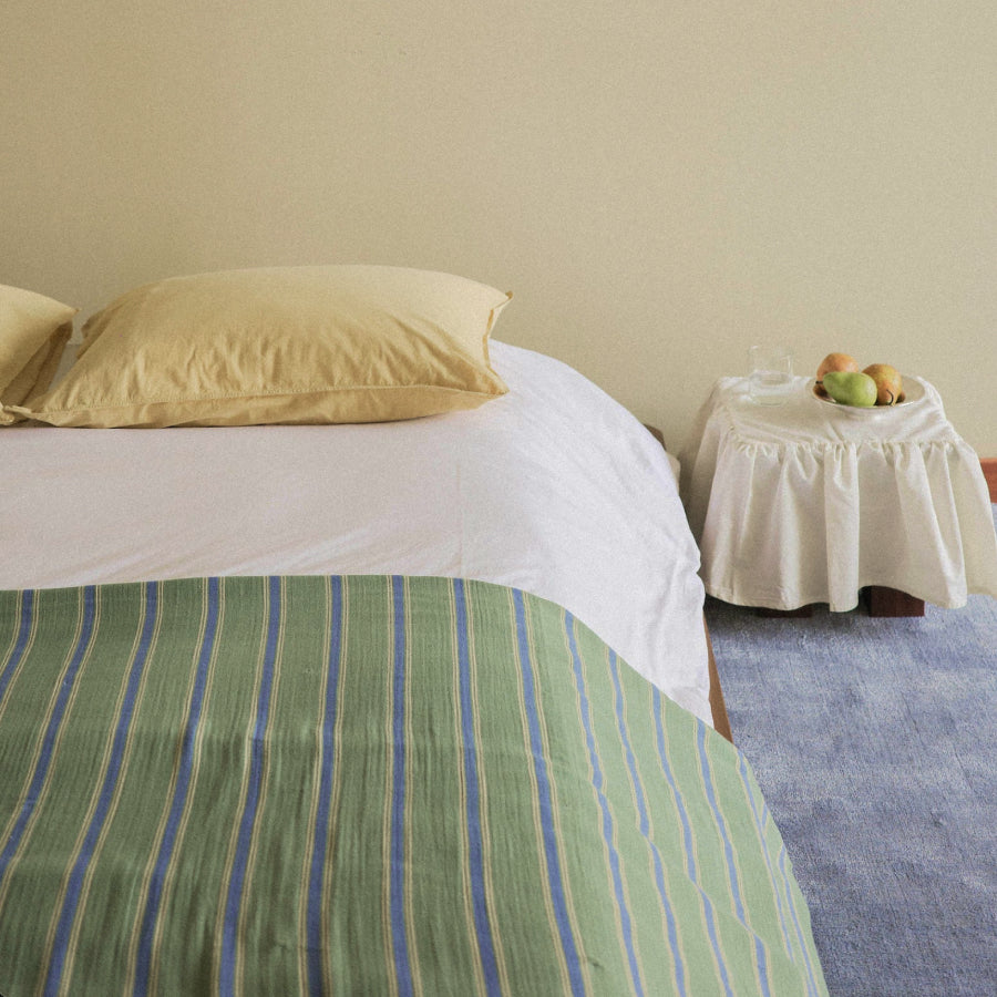A bed with white bedding and a green striped blanket, styled in a minimalist room with a cream wall, a small white-covered side table with fruit, and a blue rug.