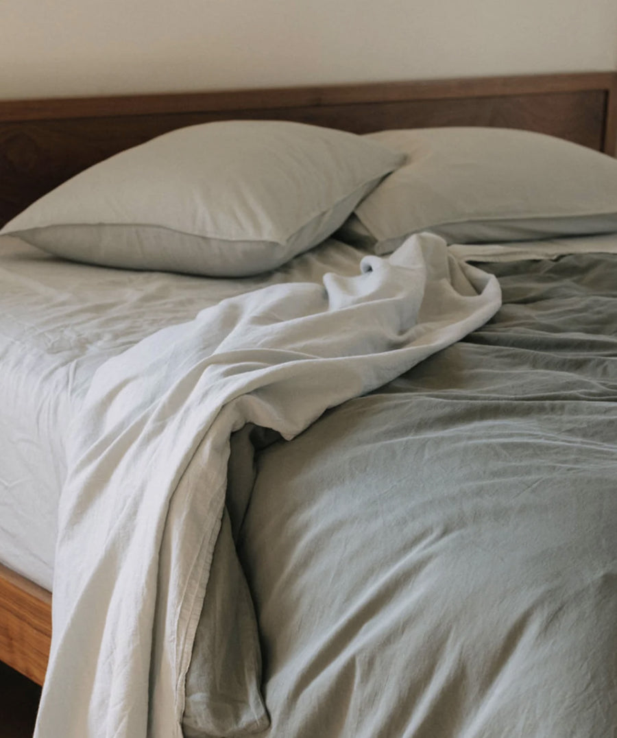 A neatly made bed with light blue bedding, including a duvet cover and two matching pillows, styled with a rumpled off-white throw blanket. The bed has a dark wood headboard and frame, set against a plain beige wall.