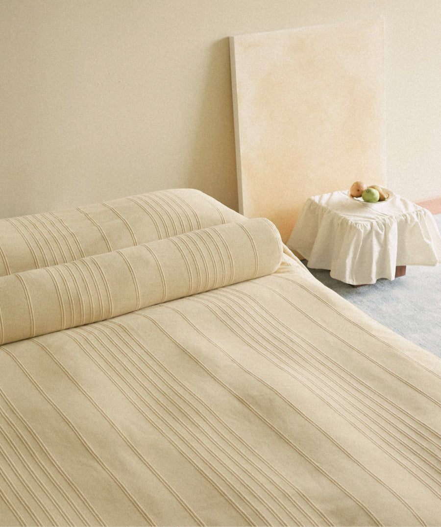 A pale yellow striped bedding set, including a duvet cover and matching pillowcases, styled on a bed in a minimalist bedroom setting with a small draped side table.