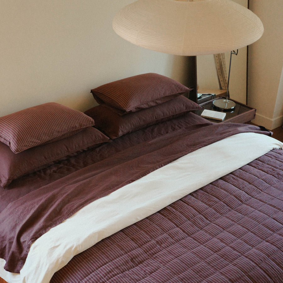 A neatly made bed in a bedroom setting, featuring a textured plum-brown quilted duvet cover, matching pillow shams, and a contrasting white folded blanket. A large woven pendant lamp hangs above, and a bedside table with a mirror and small lamp is visible against a beige wall.