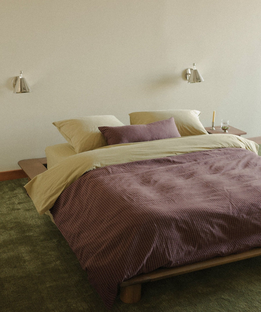 A bed dressed in pale olive and burgundy striped bedding, set in a minimalist bedroom with a wooden platform bed frame, green carpet, and wall-mounted sconces.