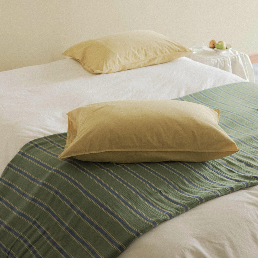 A close-up of a bed with a white duvet, green striped blanket, and a light yellow pillow in the foreground. 
