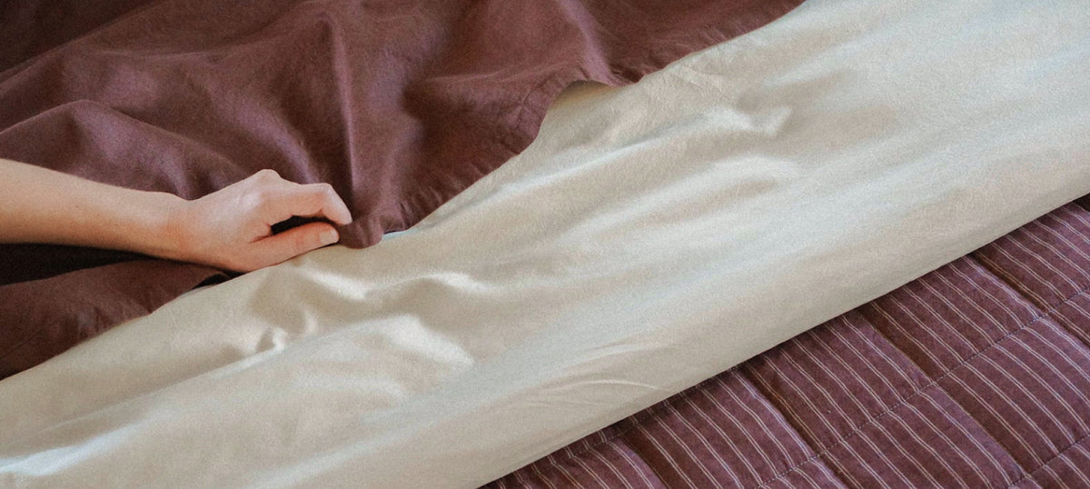 A close-up of a person's hand smoothing out a light beige sheet or duvet over a bed, with a brown striped sheet or cover underneath.