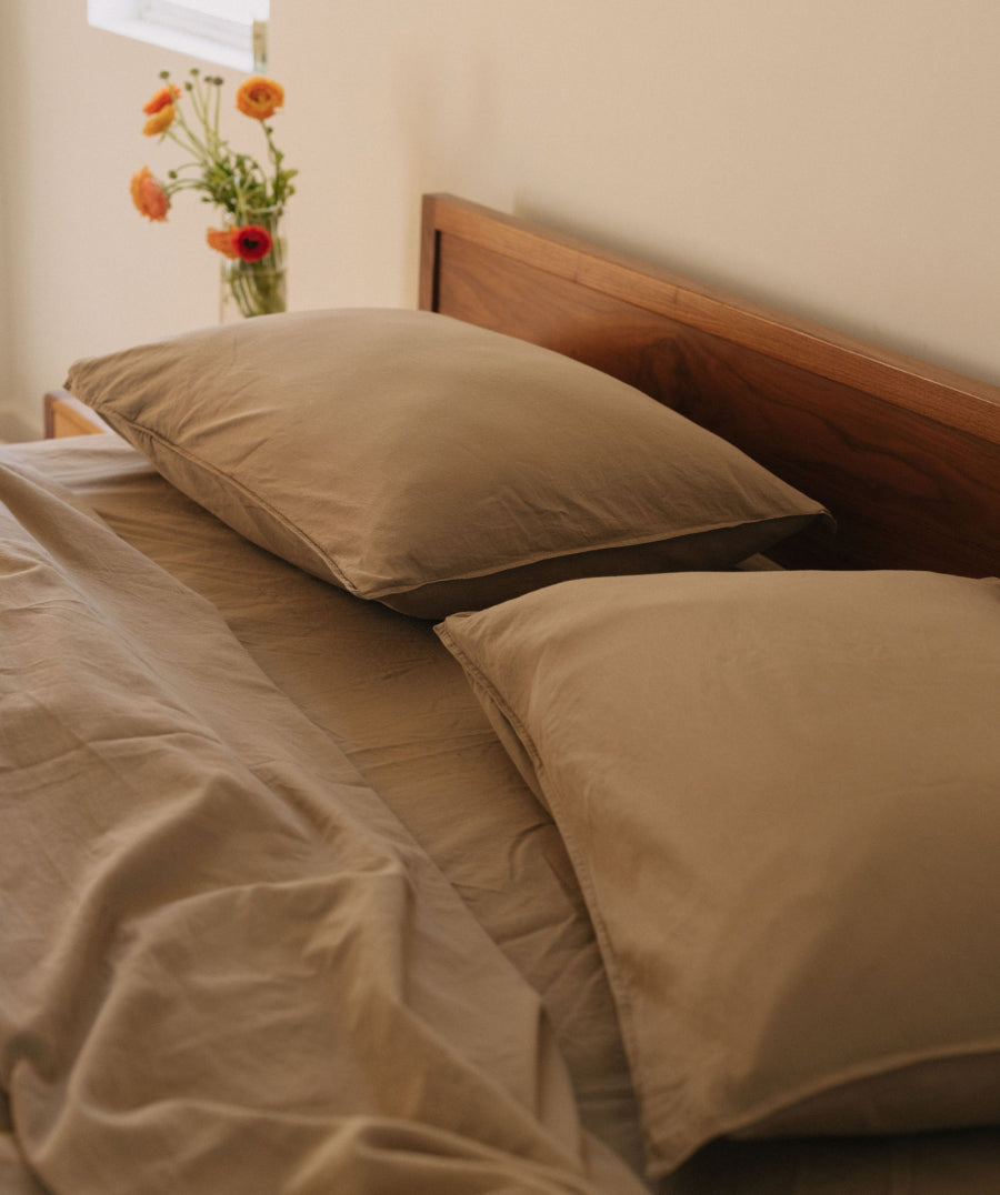 A close-up of a bed with taupe-colored sheets and two matching pillowcases, set against a dark wood headboard. A small vase of orange flowers sits on the bedside table in the softly lit background.
