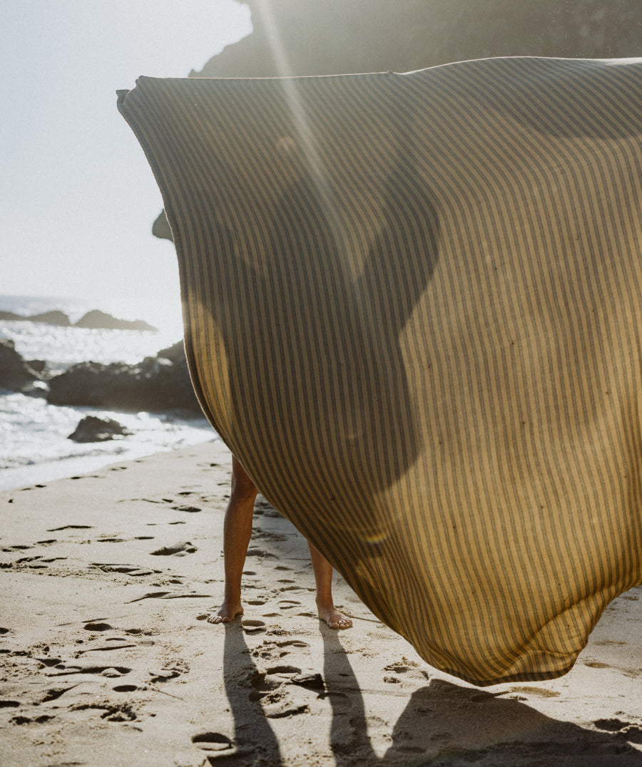 A person holding up a striped blanket standing on a sandy beach with ocean waves and rocks in the background.