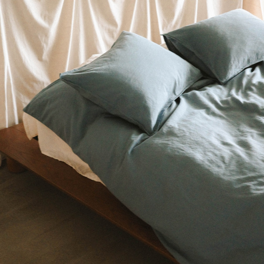 A close-up of a bed dressed in blue bedding with matching pillows, set on a wooden bed frame against a soft cream curtain backdrop.