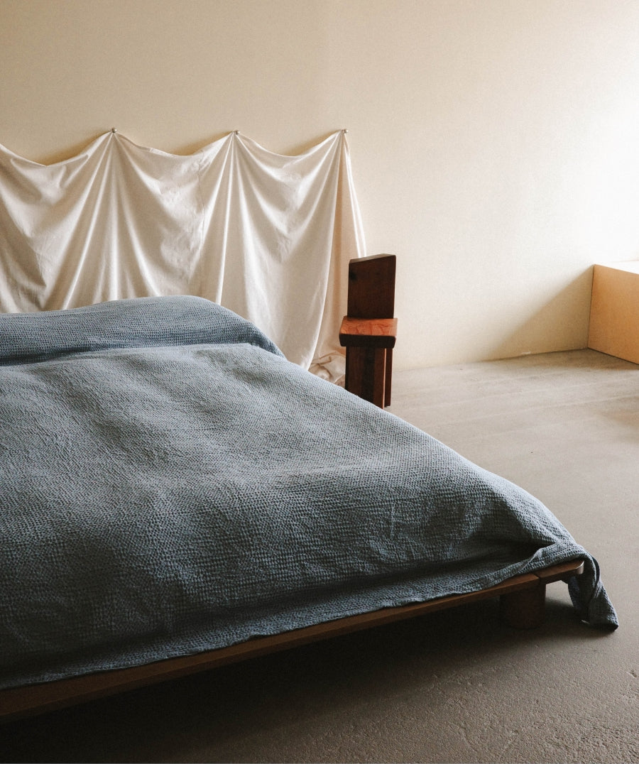 A bed dressed in a textured blue blanket and matching pillow, set in a minimalist room with a draped cream fabric headboard and a small wooden chair.