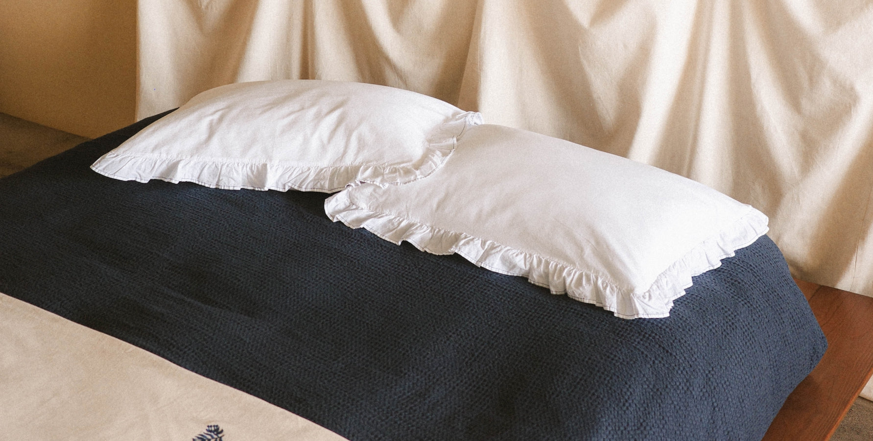 Two white ruffled pillowcases resting on a dark navy textured bedspread, set against a backdrop of draped cream fabric.