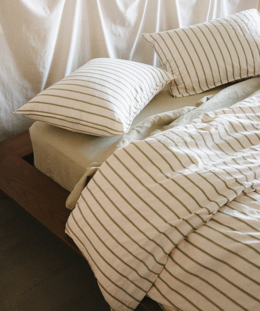 A close-up of a bed dressed in green striped bedding, including a duvet cover and two pillows, in a warm neutral bedroom setting.