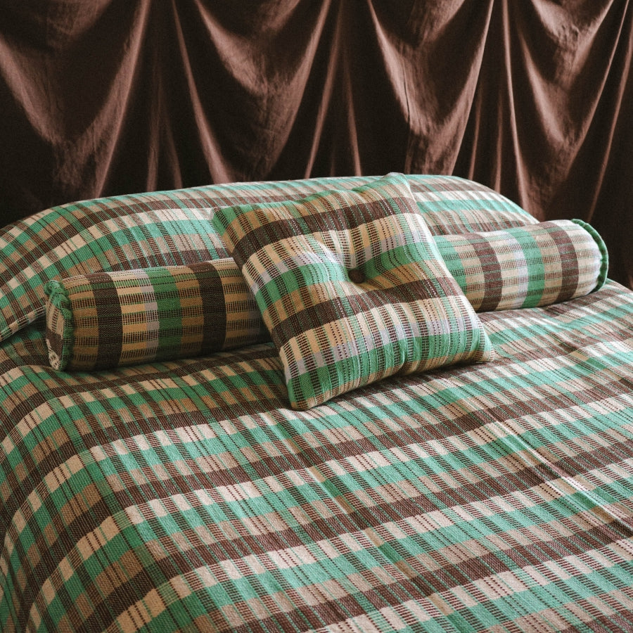 A bed dressed in a plaid bedding set with matching pillowcases and a rolled lumbar pillow. The fabric features a striped plaid pattern in green, brown, beige, and white, set against a dark brown draped curtain backdrop.