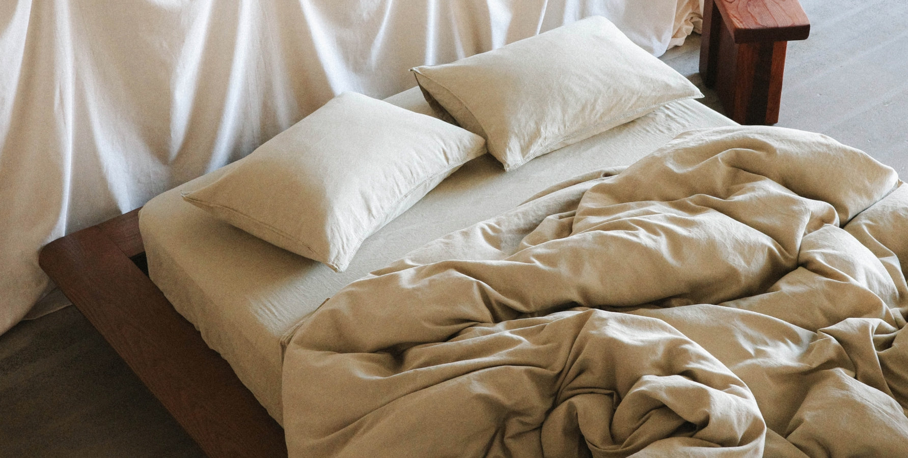 A bed dressed in light green bedding with two matching pillows and an unmade duvet, set in a bedroom with a dark wood bed frame and draped white fabric in the background.