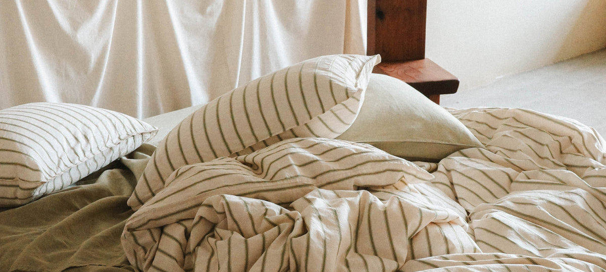 A cozy bed styled with green and white striped bedding, including a duvet cover, pillows, and a matching pillowcase, set in a softly lit bedroom with neutral-toned walls and a wooden headboard partially visible.