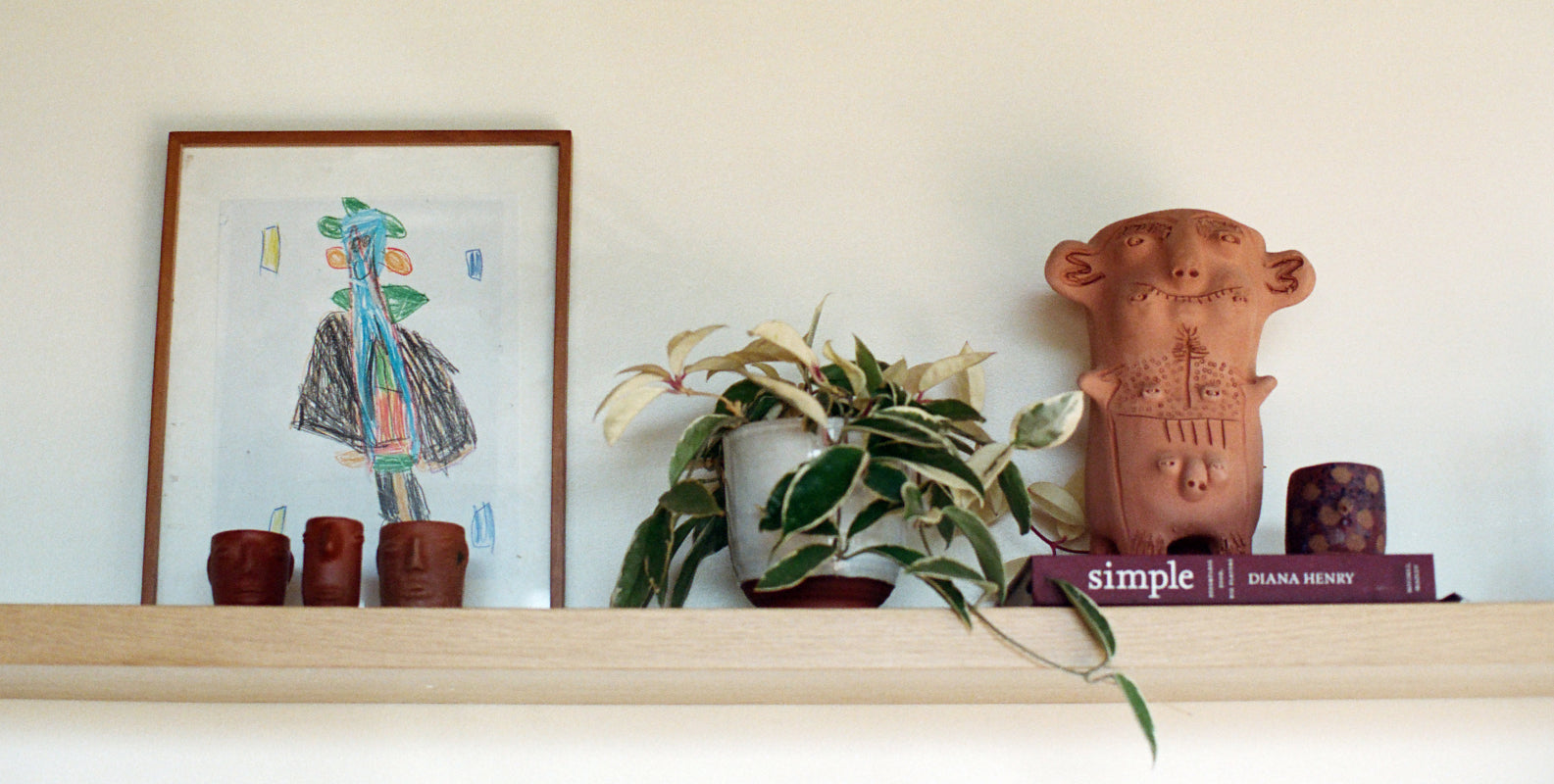 A terracotta-colored ceramic monkey figurine with carved details sits on a wooden shelf next to a potted variegated plant and a framed abstract drawing on a white wall.
