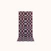 A long rectangular woven mat featuring a geometric checkerboard pattern in light blue, red and plum set against a solid off-white background. 