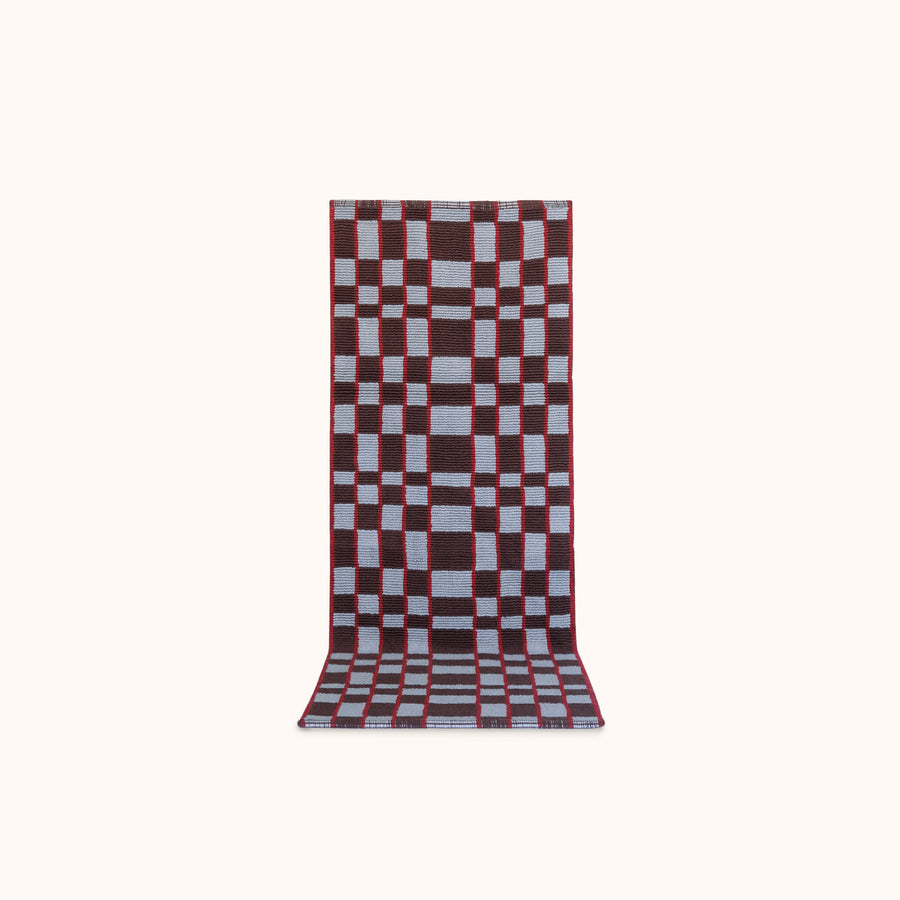 A long rectangular woven mat featuring a geometric checkerboard pattern in light blue, red and plum set against a solid off-white background. 