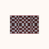 A rectangular woven mat featuring a geometric checkerboard pattern in light blue, red and plum set against a solid off-white background. 