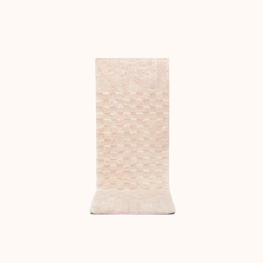 A rectangular, blush-colored bath mat with a subtle check pattern and contrasting shag heights.