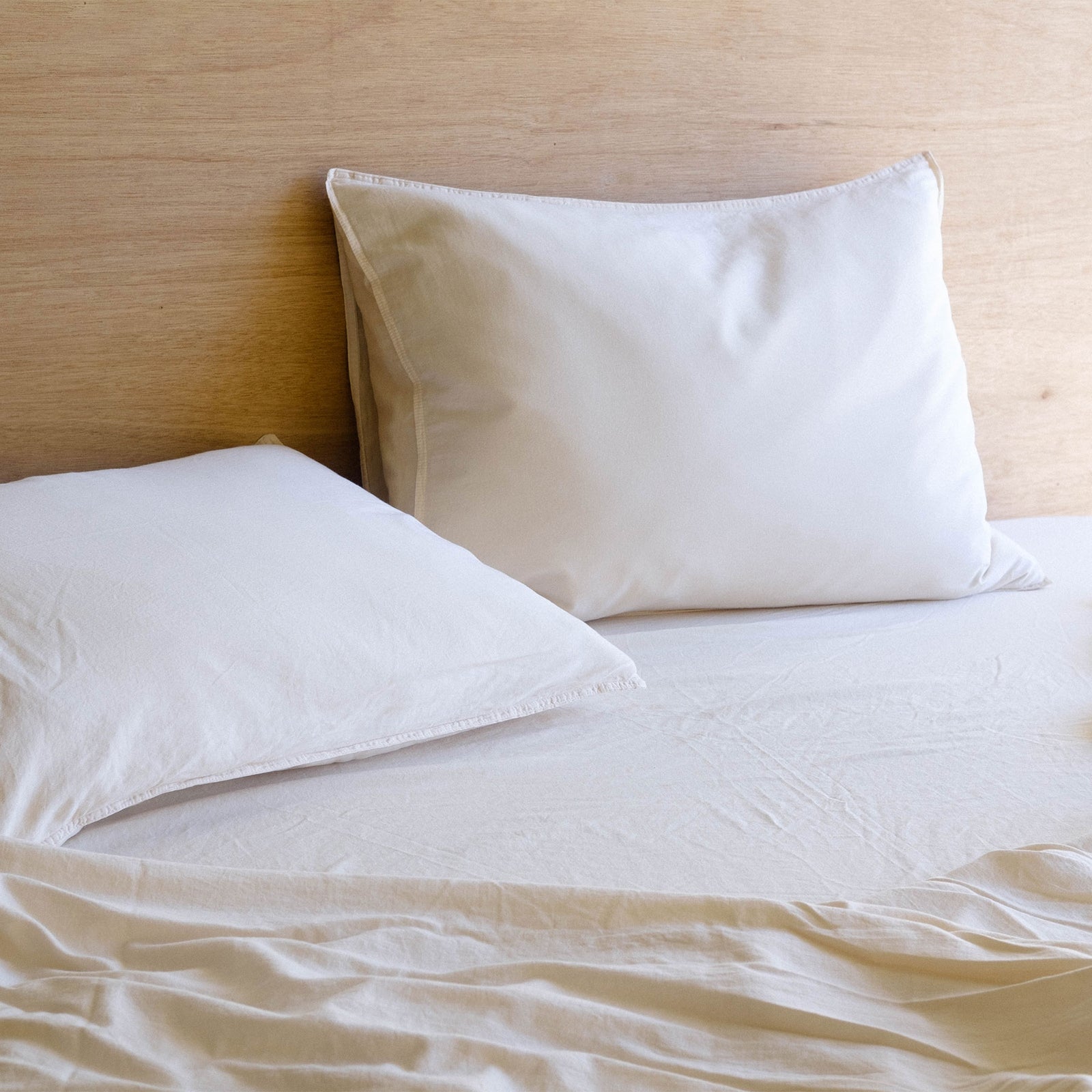 Two pillows on a bed - the pillows and sheets are Oat / cream colored and the pillows are against a large wooden headboard