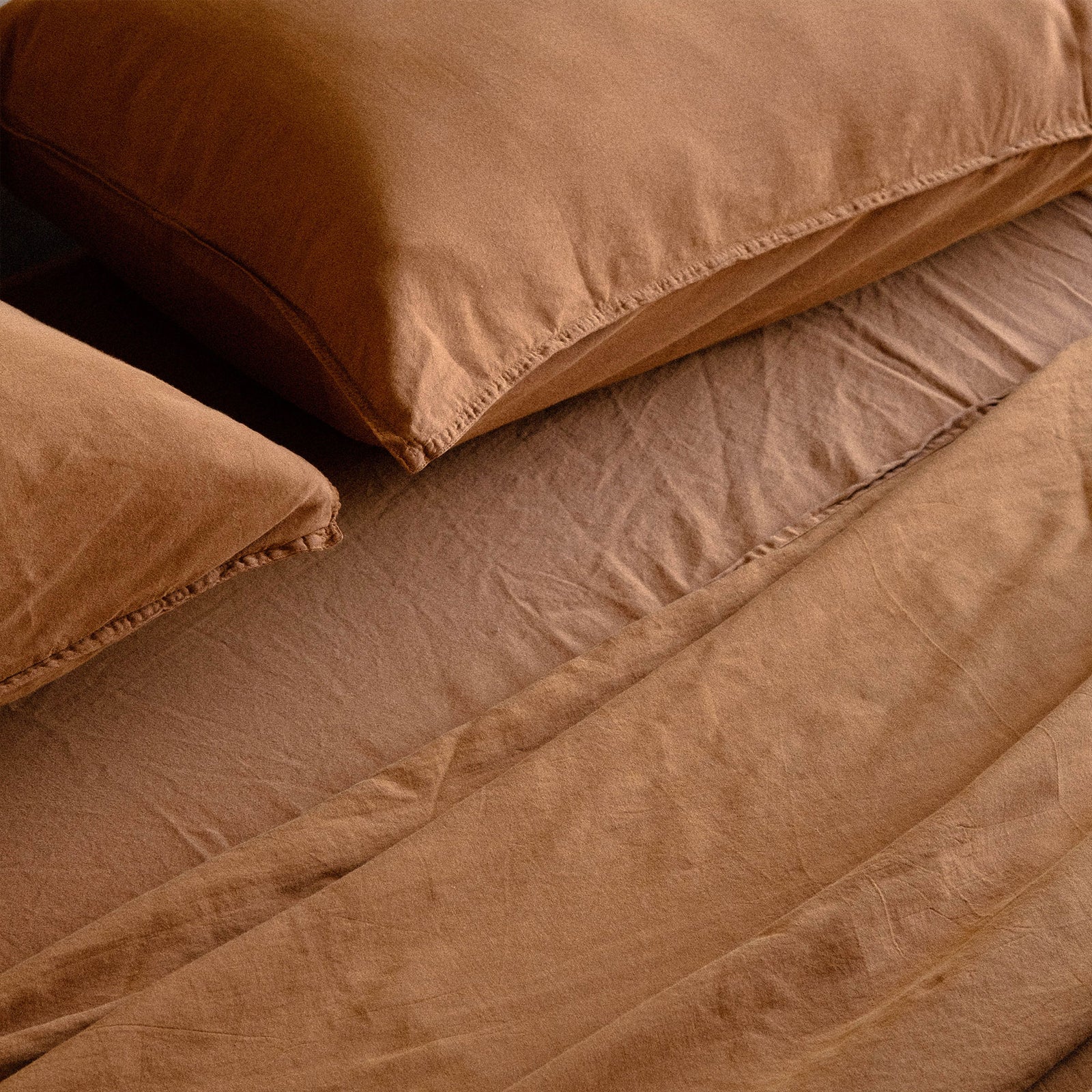 A details shot of the edge of two cotton matte sateen pillows in terracotta / brown on a bed with matching sheets
