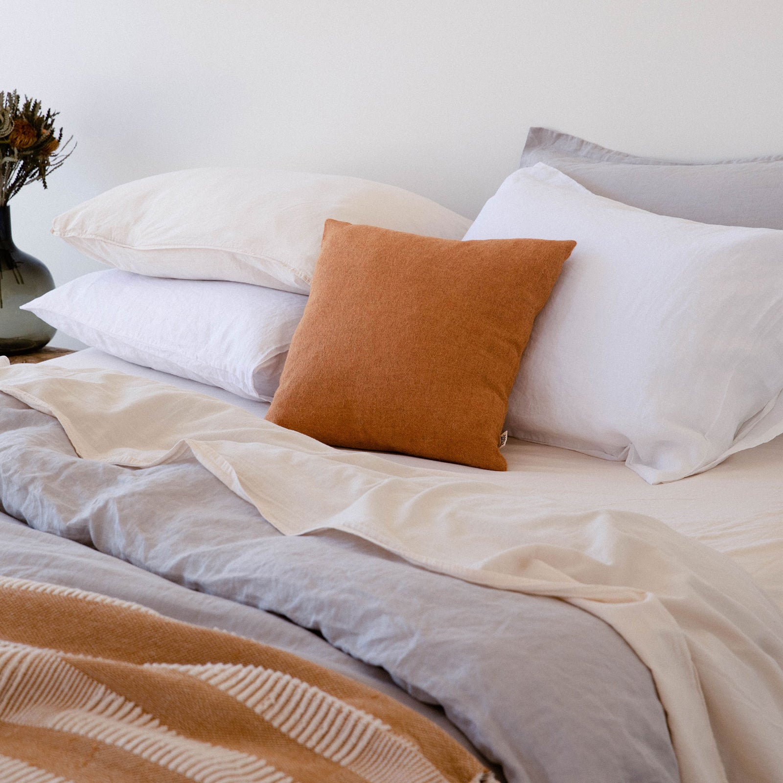 Oat / cream and white organic matte sateen pillows stacked and laying on a bed with grey sheets and a tan throw. There is a tan throw pillow at the center. 