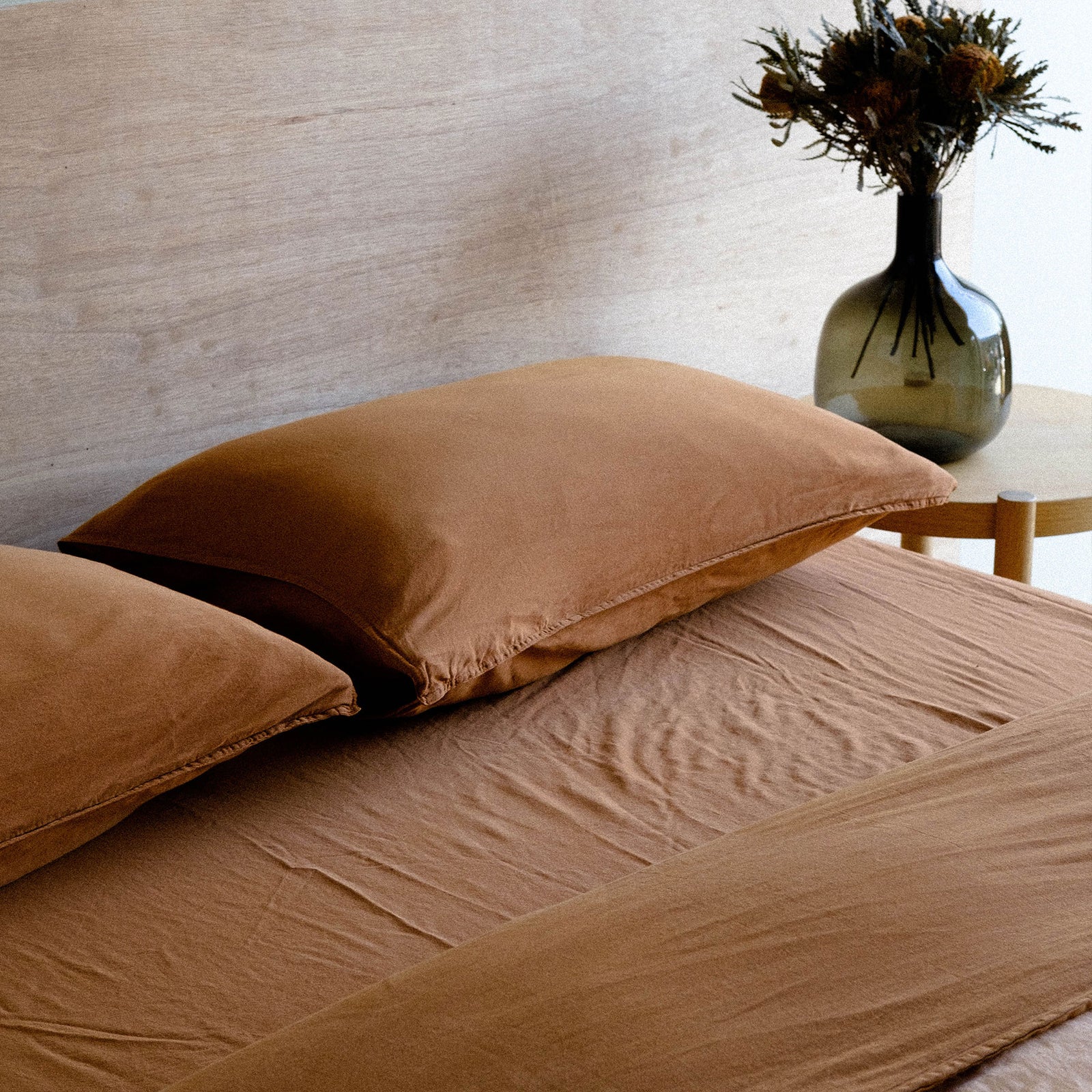 A neatly made bed with matte sateen sheets in terracotta in a bedroom setting with a vase of dried flowers in the background