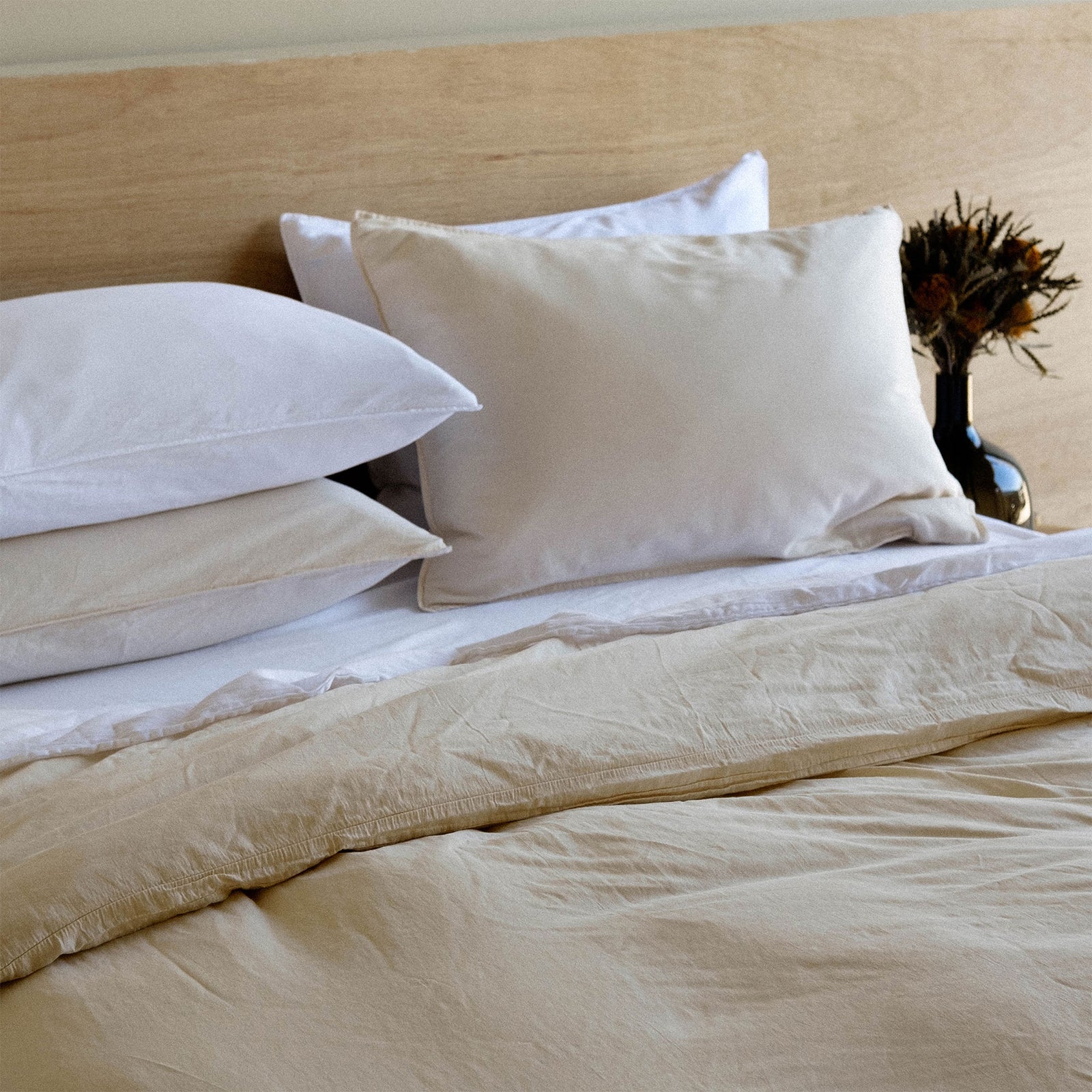 A slightly different angle of a bed made with white, oat and beige sheets with a vase of dried flowers in the background