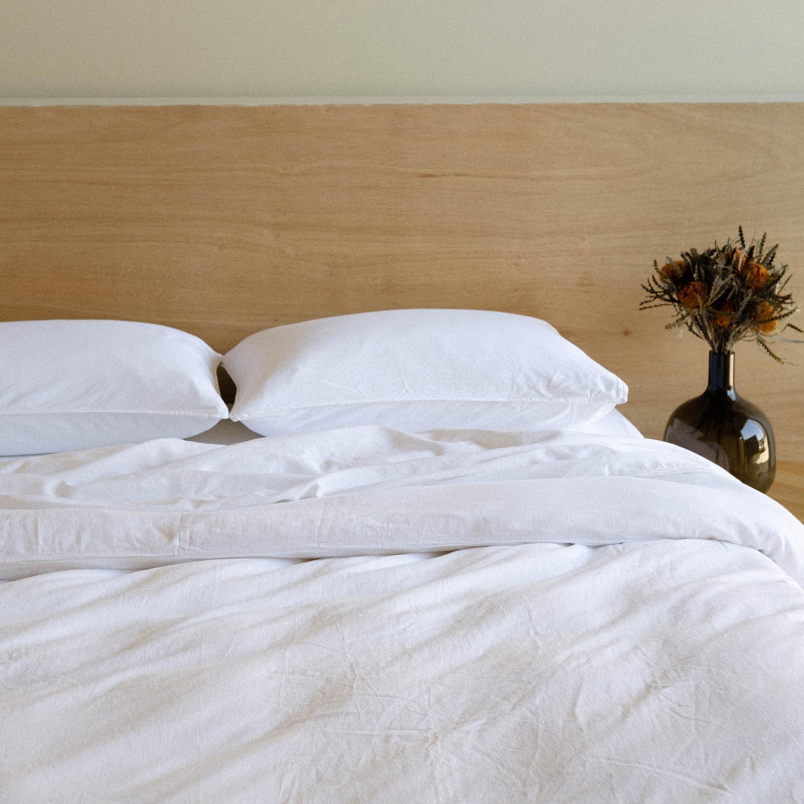A neatly made bed of white matte sateen sheets against a wooded headboard. There is a base with dried flowers beside the bed. 